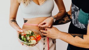 A couple measuring waistline with a tape and holding a fresh salad bowl, focusing on fitness and weight loss.
